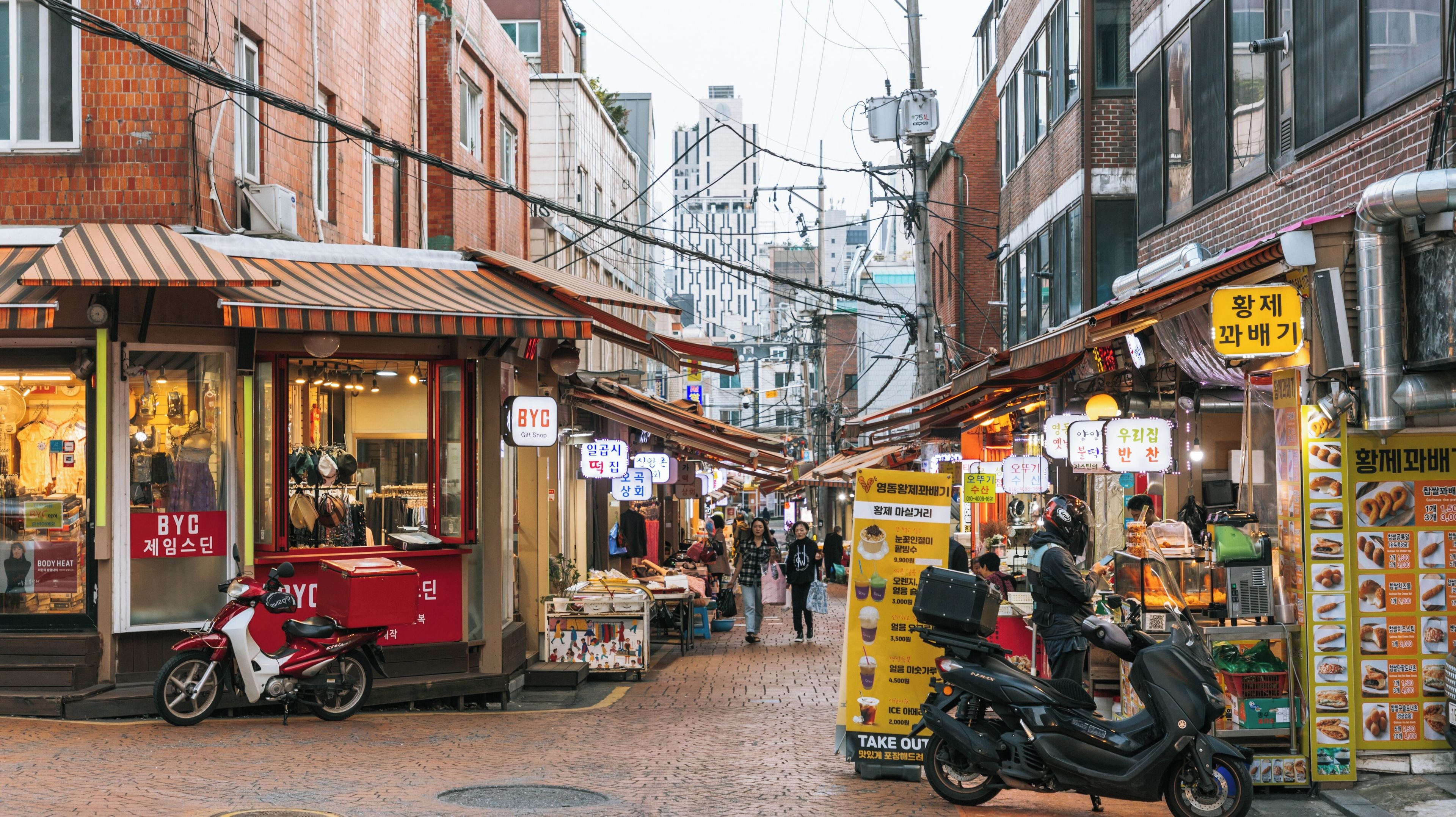 Rua estreita com lojas, uma moto vermelha e uma placa amarela.