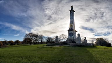 Un monument composé d’une haute colonne surmontée d’une sphère, entouré d’un champ verdoyant et d’arbres, sous un ciel partiellement nuageux.