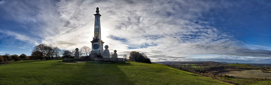 A monument with a tall column and spherical top, surrounded by a grassy field and trees, under a partly cloudy sky.