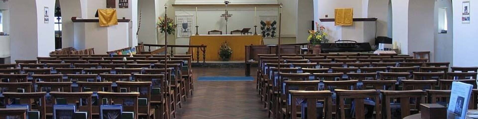 An interior of a church with rows of wooden pews, a central altar, and a stained glass window.