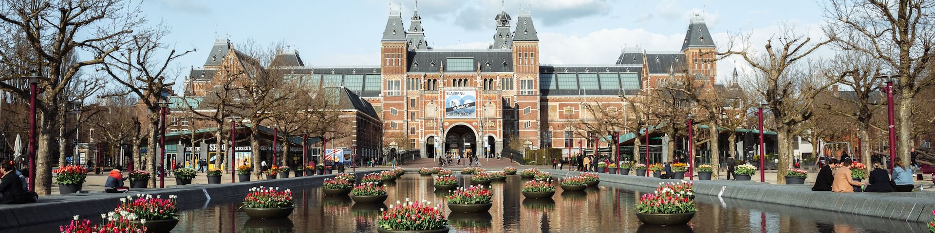 A historic building with a large central archway, surrounded by a reflective water feature with flower planters.