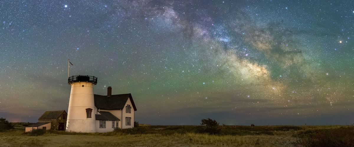 A lighthouse with a house nearby, under a starry night sky with visible Milky Way.