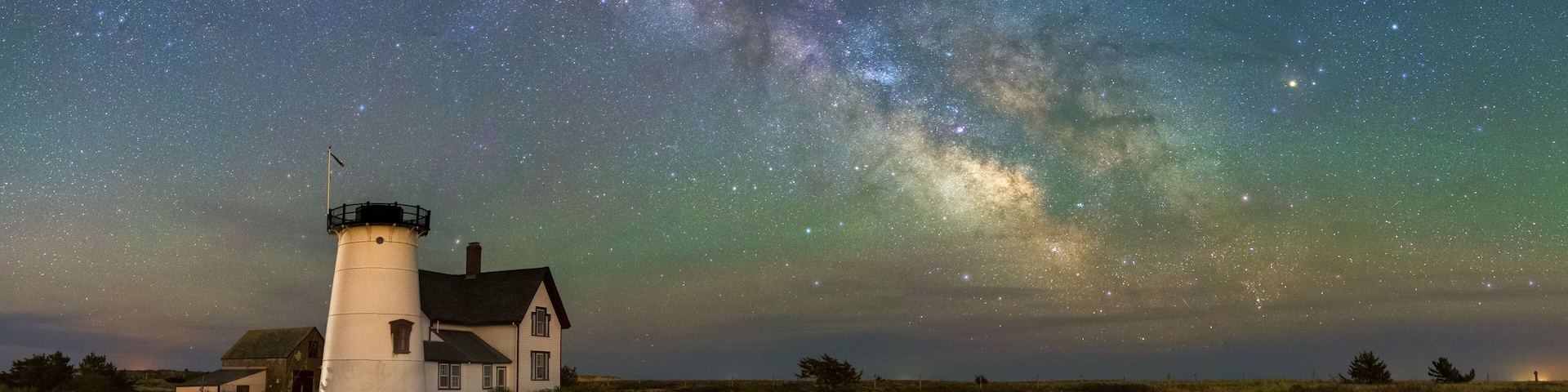 A lighthouse with a house nearby, under a starry night sky with visible Milky Way.