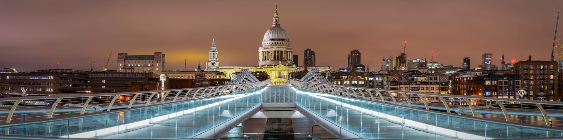 Uma ponte moderna com painéis de vidro, que conduz a um edifício histórico com cúpula e torre do relógio, com o horizonte da cidade ao fundo ao entardecer.