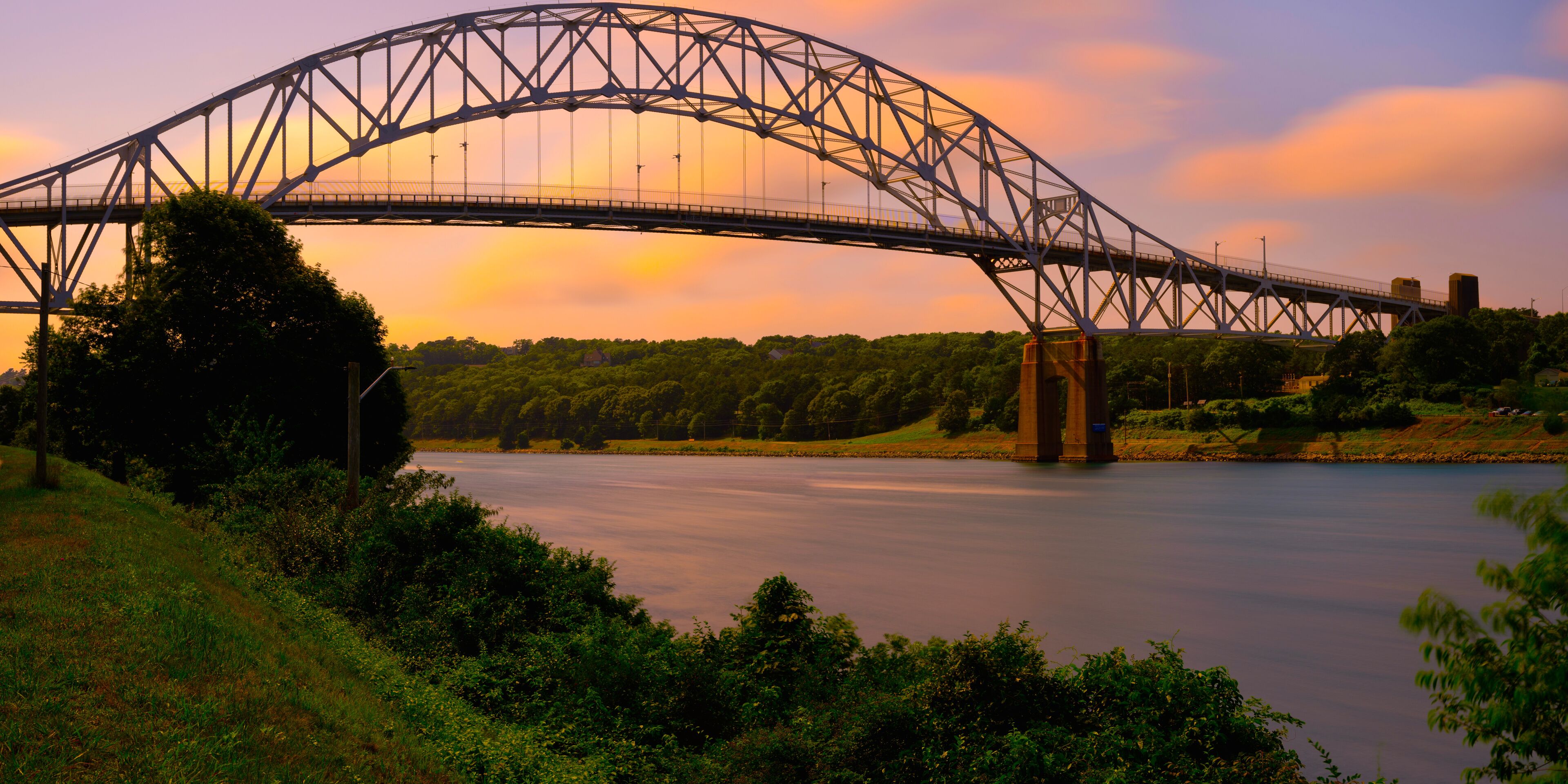 Sagamore Bridge at Sunrise over Cape Cod Canal with floating orange colored clouds and green bushes and tree on the riverbank on Cape Cod, Massachusetts, USA, Long Exposure photo