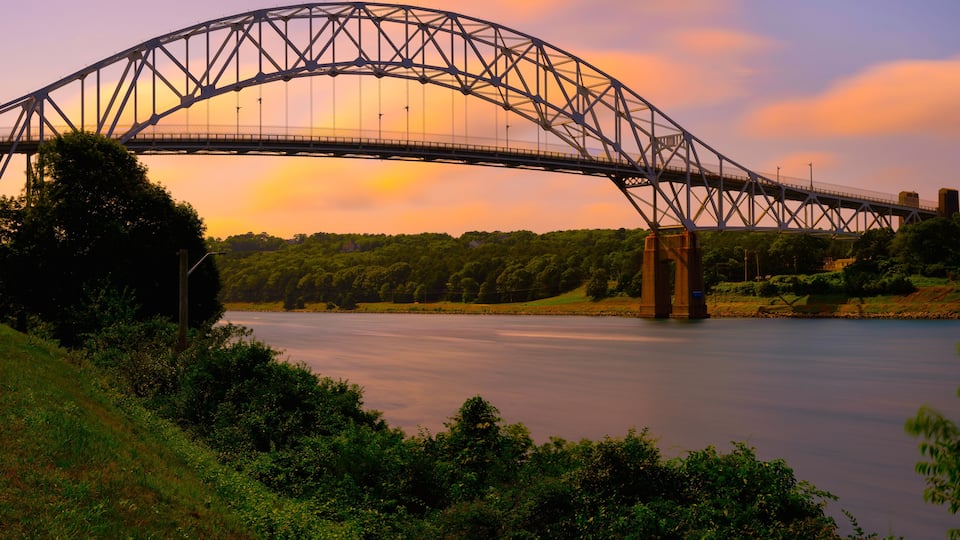 Sagamore Bridge at Sunrise over Cape Cod Canal with floating orange colored clouds and green bushes and tree on the riverbank on Cape Cod, Massachusetts, USA, Long Exposure photo