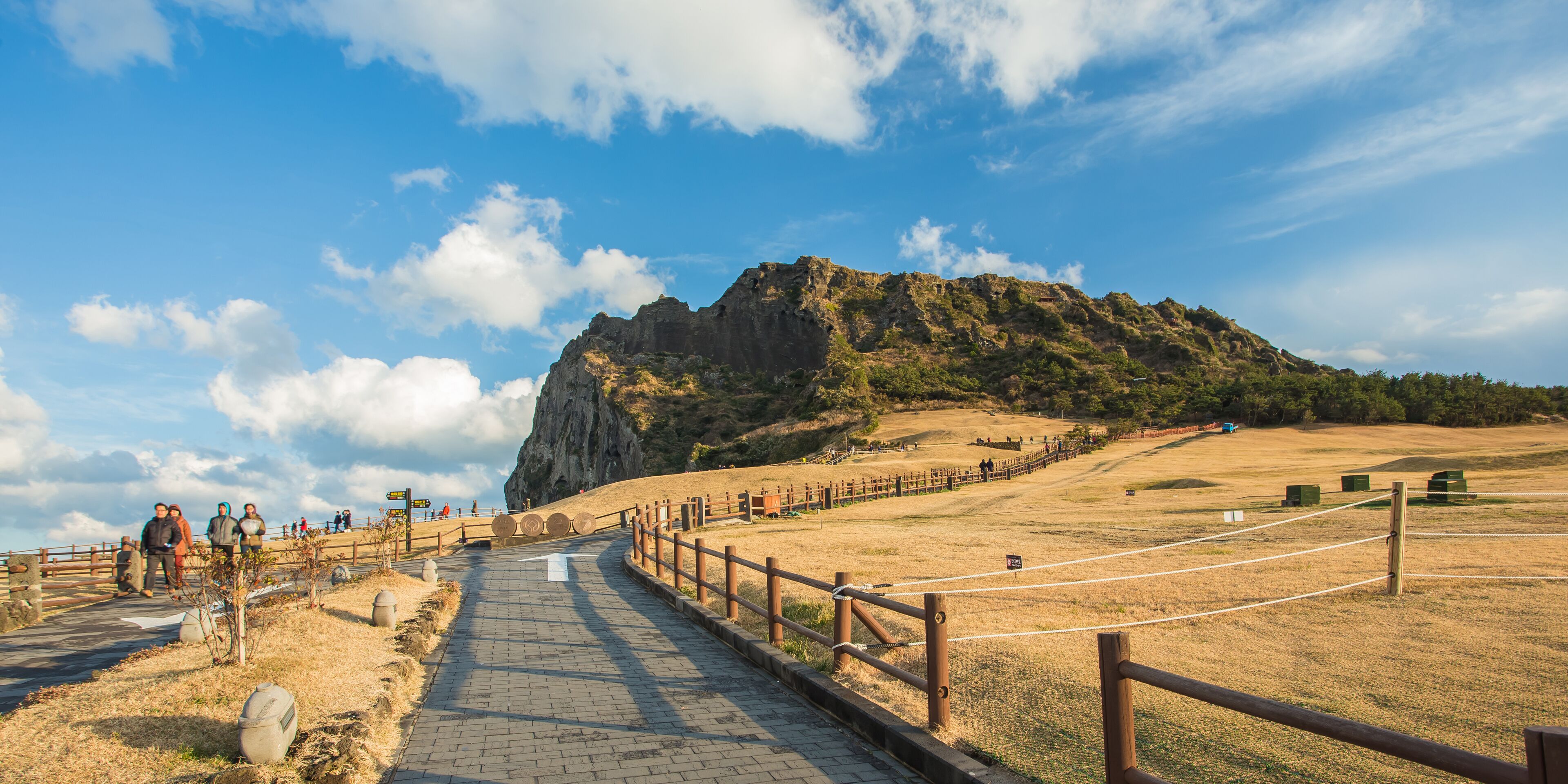 A paved pathway leading to a rocky hill with a wooden fence and a few people walking.