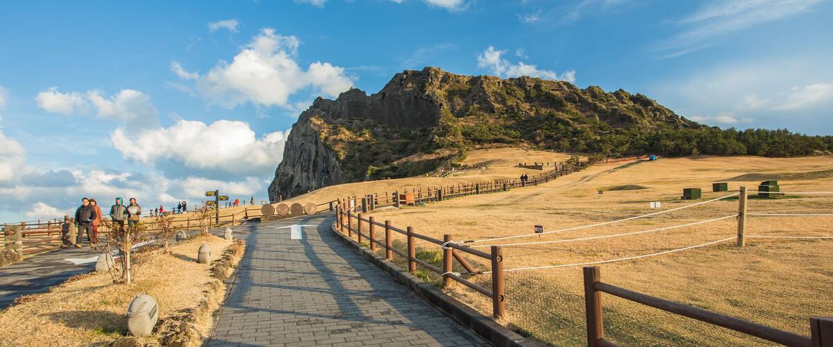 A paved pathway leading to a rocky hill with a wooden fence and a few people walking.
