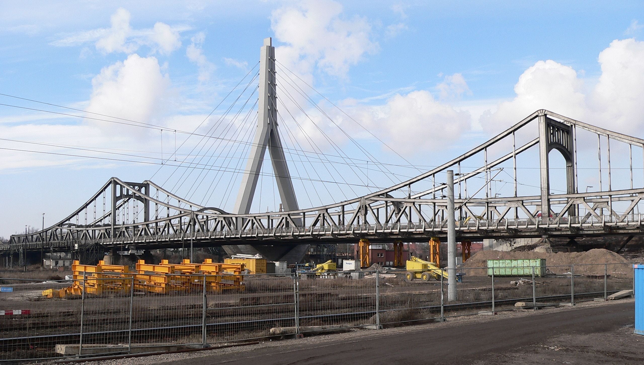 A bridge with a tall central tower and cables, construction equipment, and a fenced area.