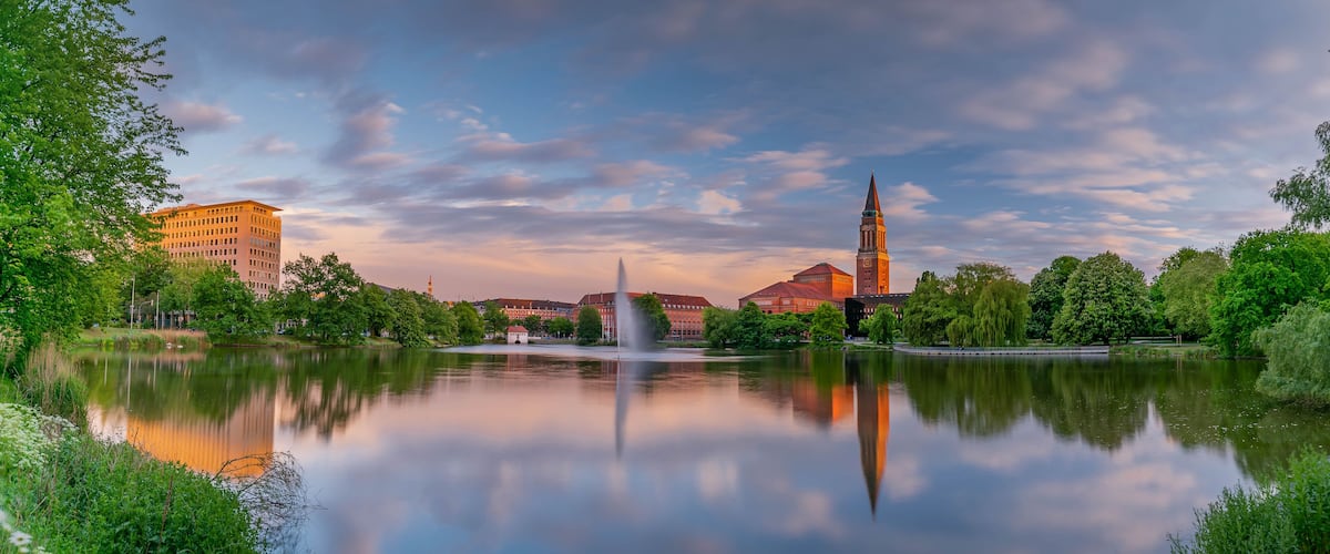 Ein friedlicher See mit dem Spiegelbild eines hohen Turms und eines Gebäudes, umgeben von üppigem Grün und blauem Himmel.