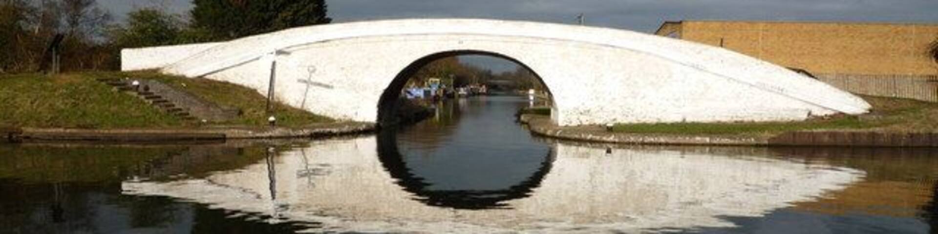 A white stone bridge over a calm river with a reflection, surrounded by trees and a cloudy sky.