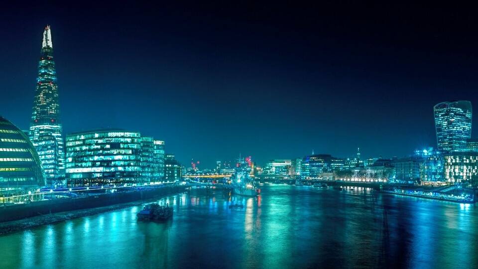 A nighttime cityscape with illuminated buildings and a river reflecting the lights.