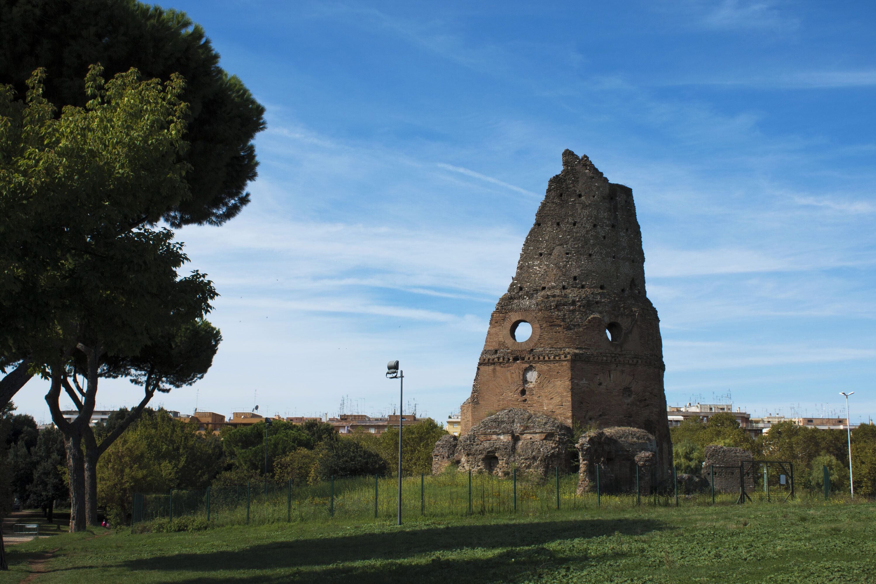 Una torre histórica con un ventanal circular, rodeada de vegetación y edificios urbanos al fondo.