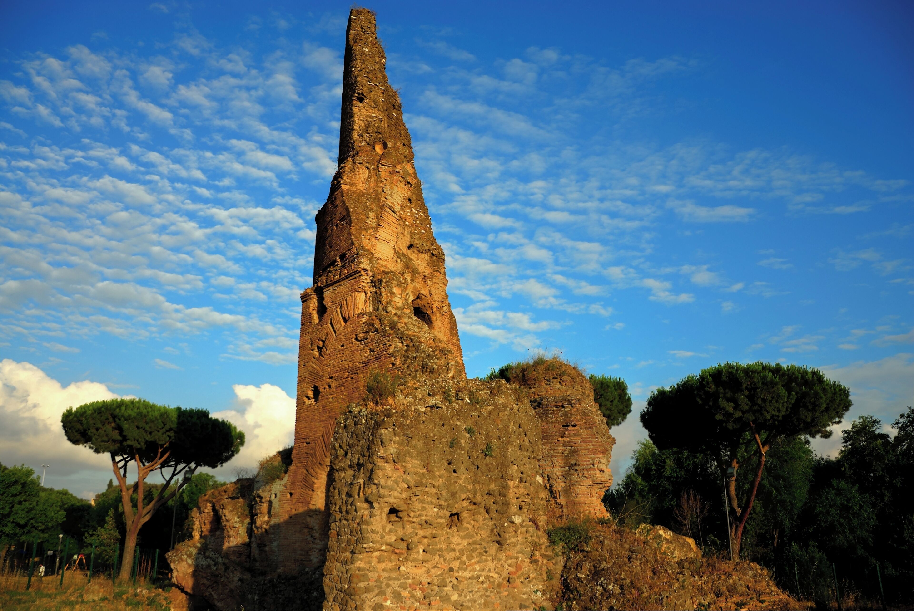 Una alta y antigua torre de piedra con un techo cónico, rodeada de árboles y ruinas.