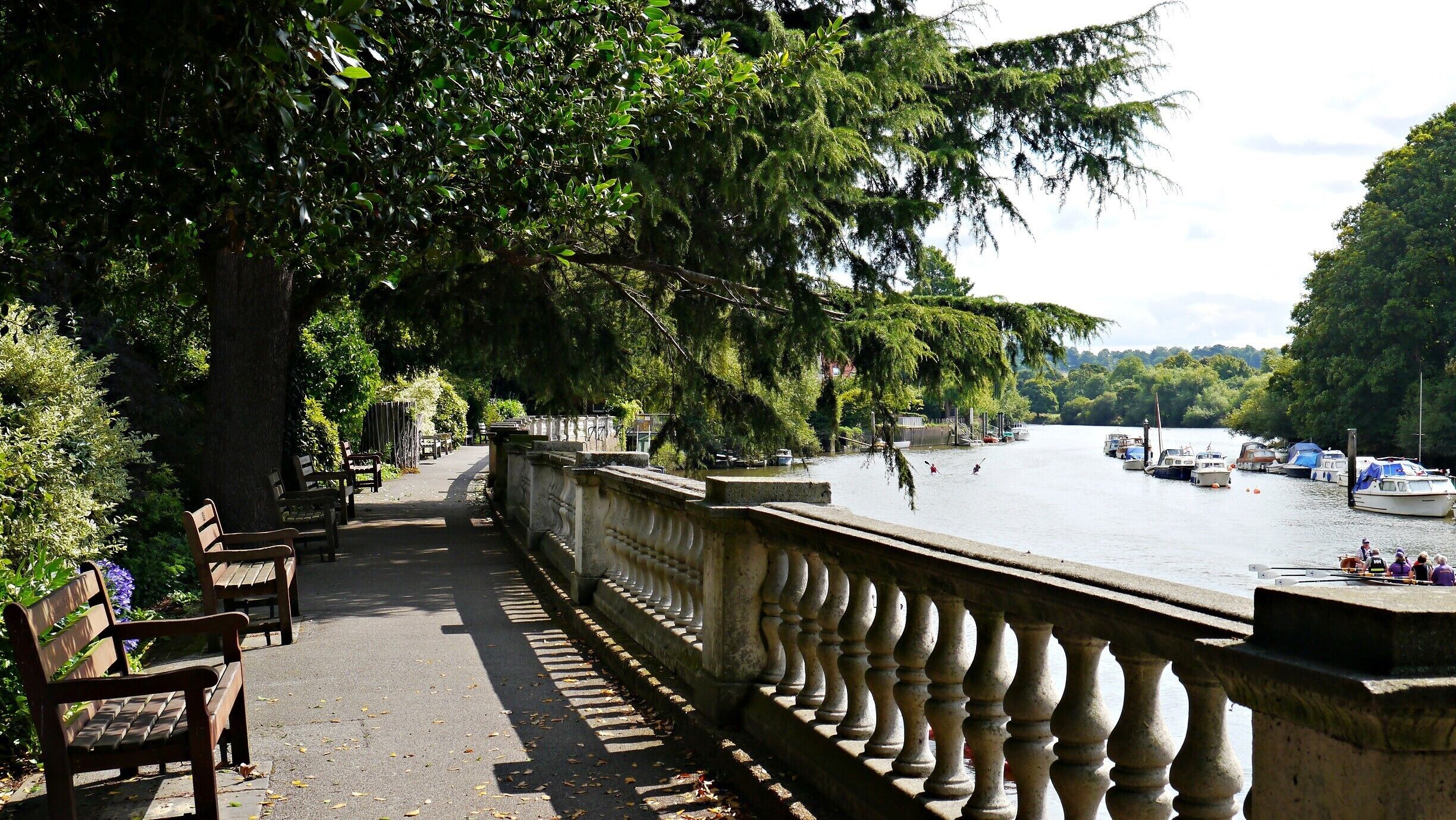 A riverside walkway with benches, a stone balustrade, and boats on the water.