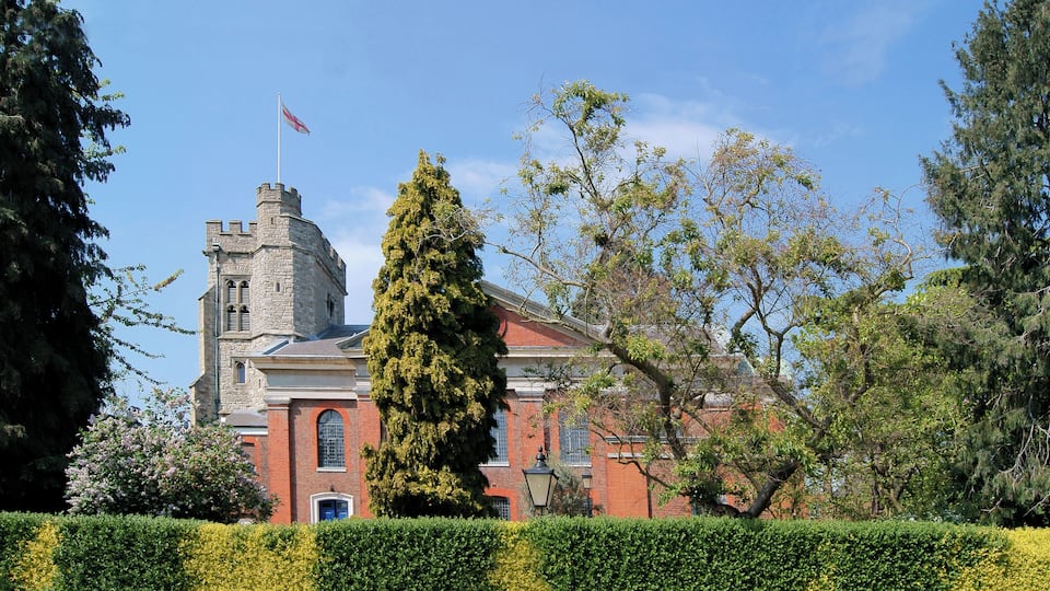 A historic building with a tower and flag, surrounded by well-maintained hedges and trees.