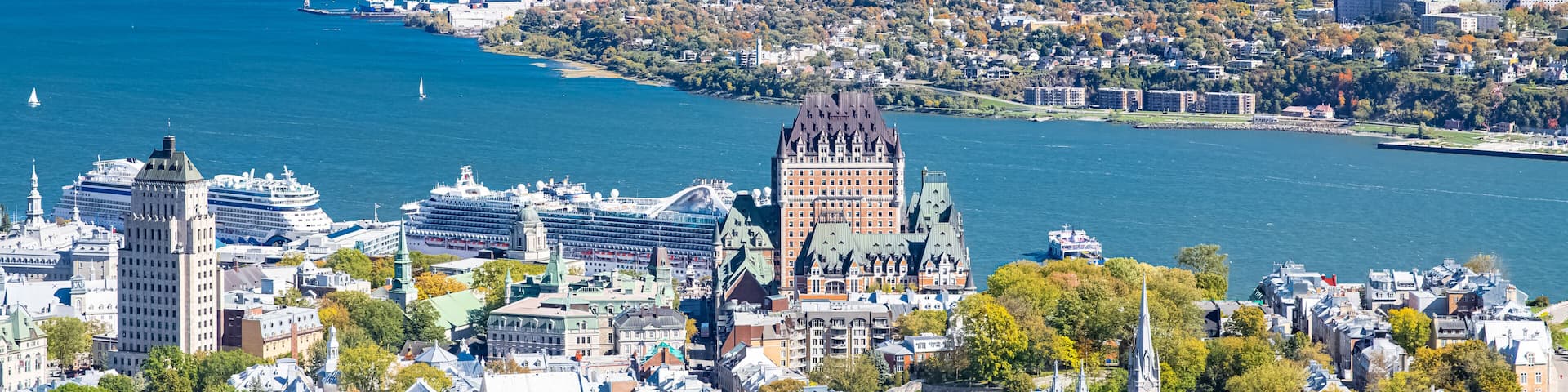 A coastal cityscape with a prominent historic building, a large cruise ship docked, and a hilly landscape in the background.