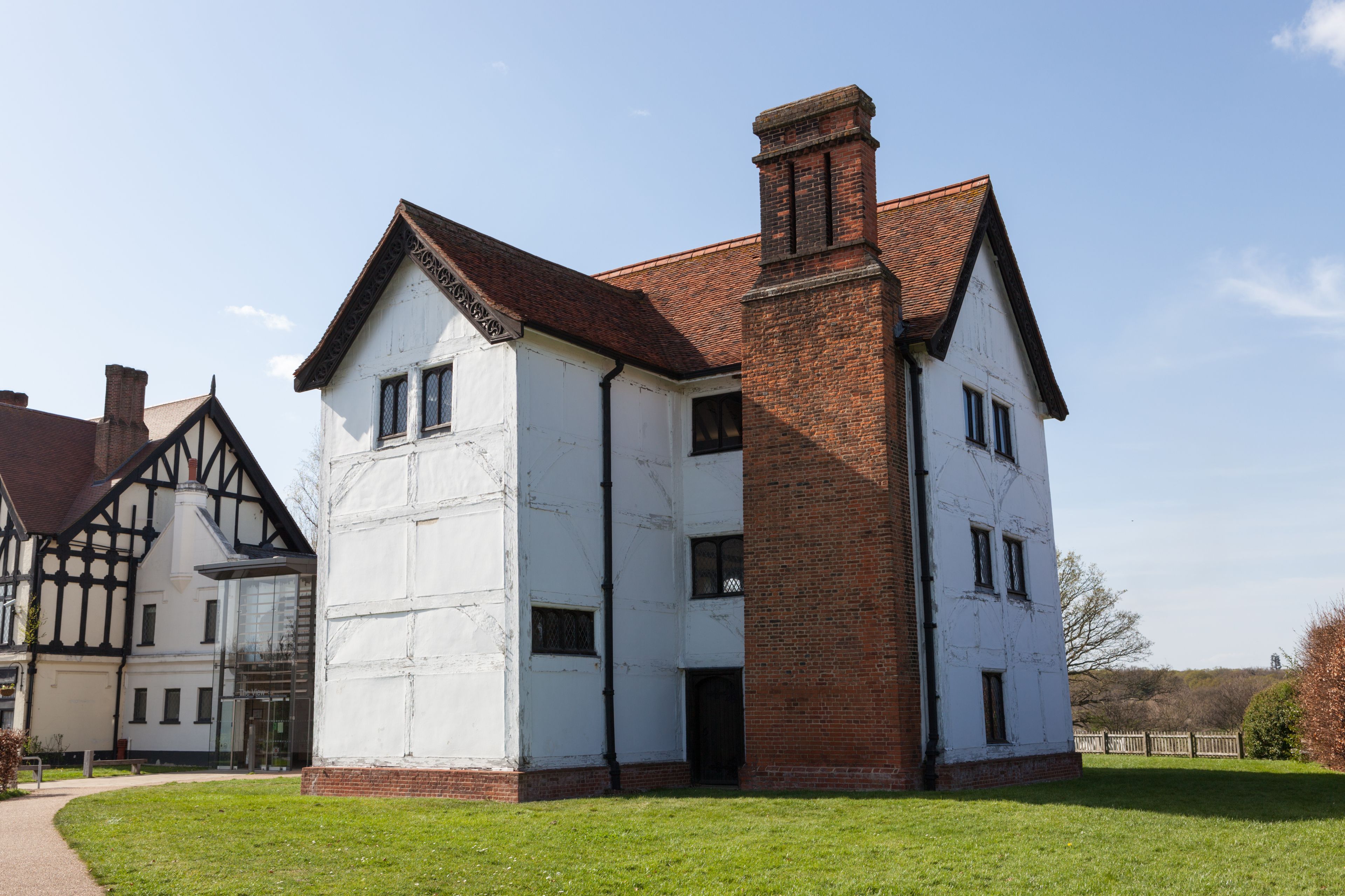 Un grande edificio bianco con un camino in mattoni rossi, circondato da un'area verdeggiante e un edificio più piccolo in stile Tudor.