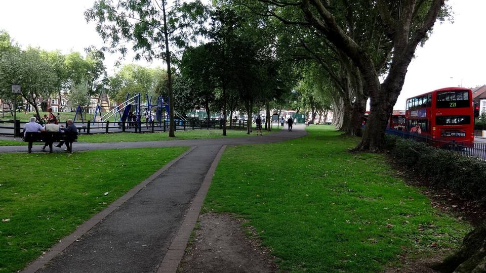 A park with a walking path, benches, playground equipment, and a red double-decker bus.