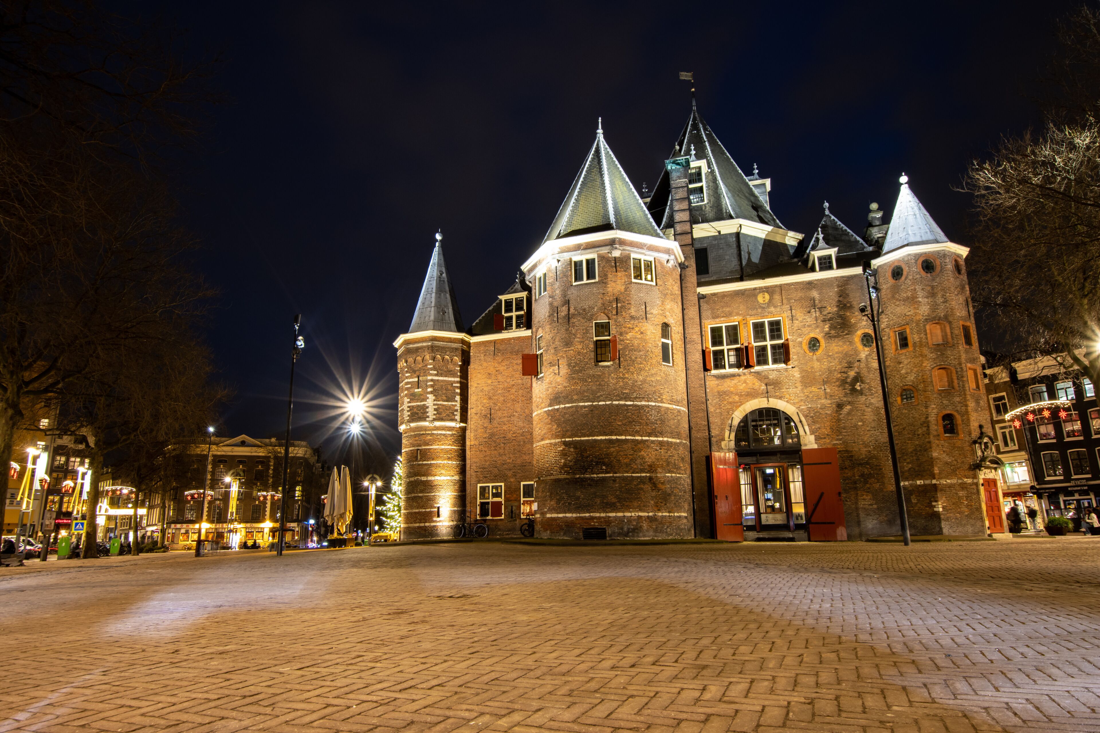 A historic castle with multiple towers and a large entrance gate, surrounded by cobblestone streets and illuminated by street lamps.
