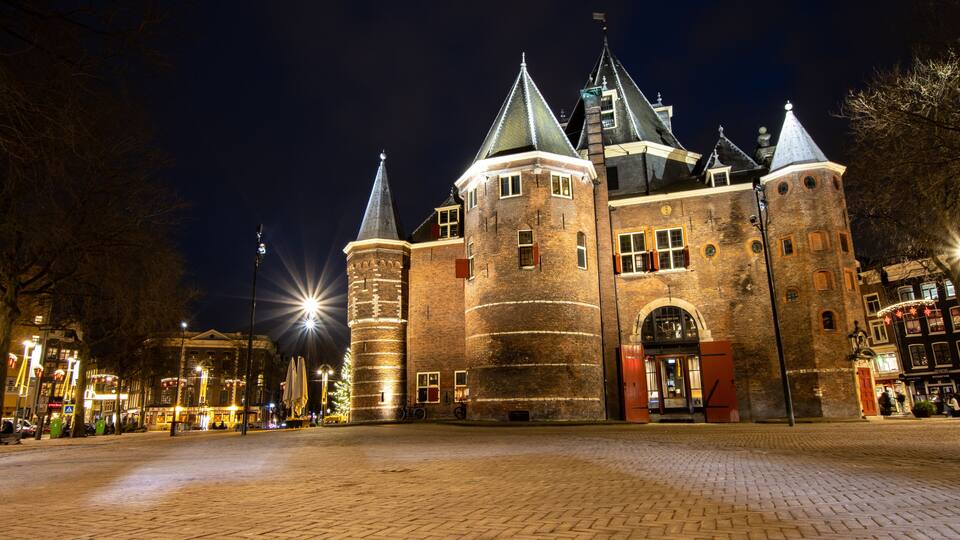 Ett historiskt slott med flera torn och en stor port, omgivet av kullerstensgator och upplyst av gatlyktor.