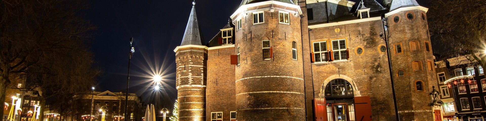 Um castelo histórico com várias torres e um grande portão de entrada, rodeado por ruas de paralelepípedos e iluminado por postes de luz.
