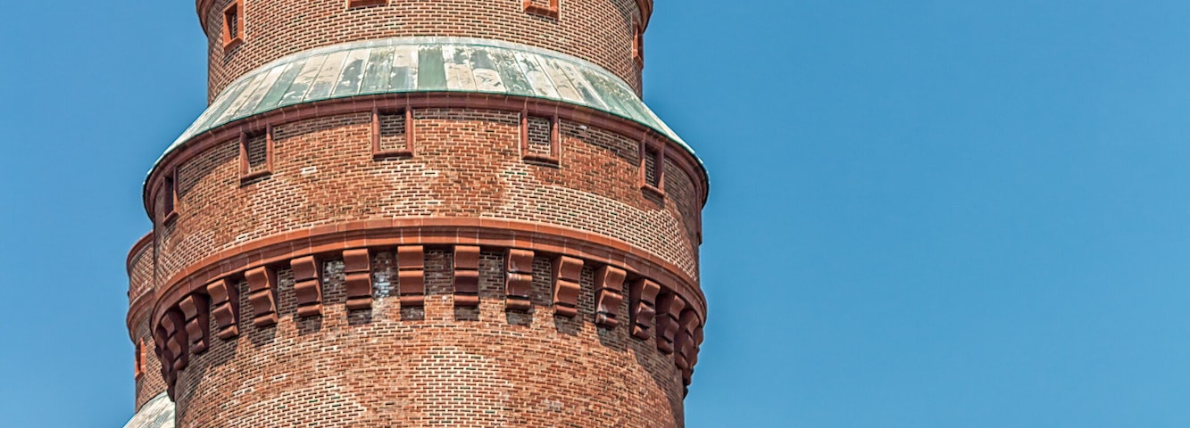 Ein hoher Backstein-Turm mit spitz zulaufendem Dach vor blauem Himmel.