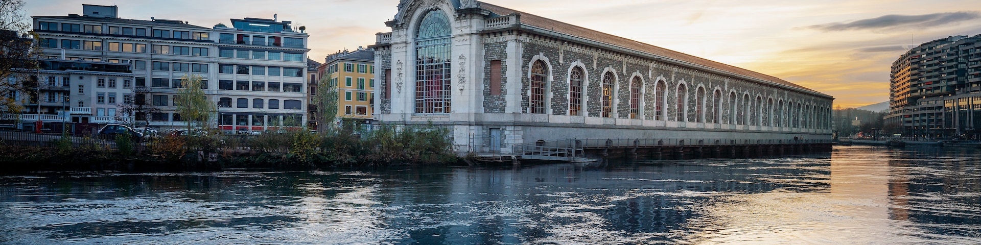 Un bâtiment historique doté d’arches et d’une coupole centrale, qui se reflète dans les eaux calmes d’une rivière, avec des bâtiments modernes en arrière-plan.