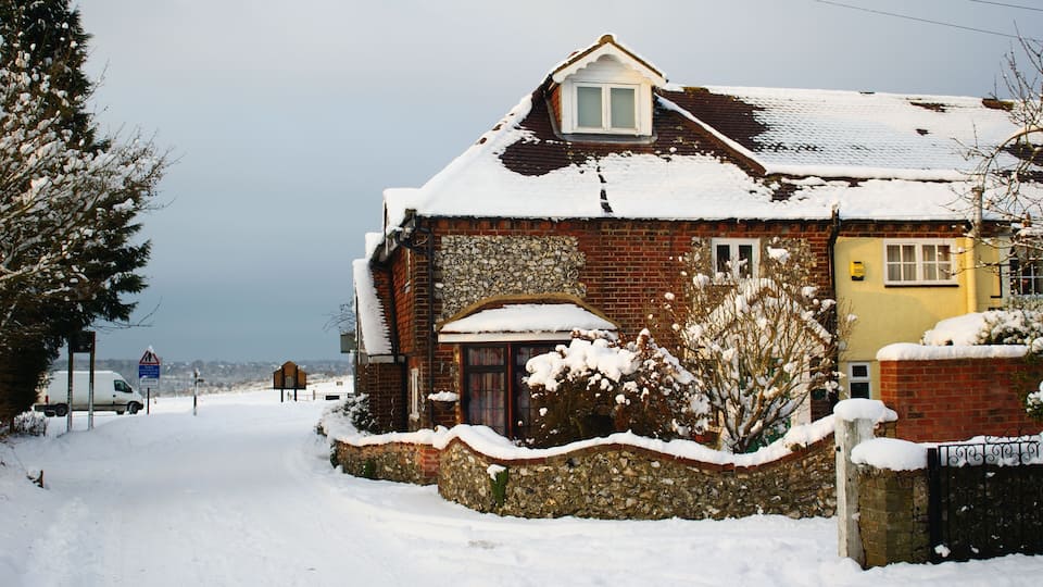Una casa cubierta de nieve con un muro de piedra y fachada de ladrillo, rodeada de árboles nevados y un cielo despejado.