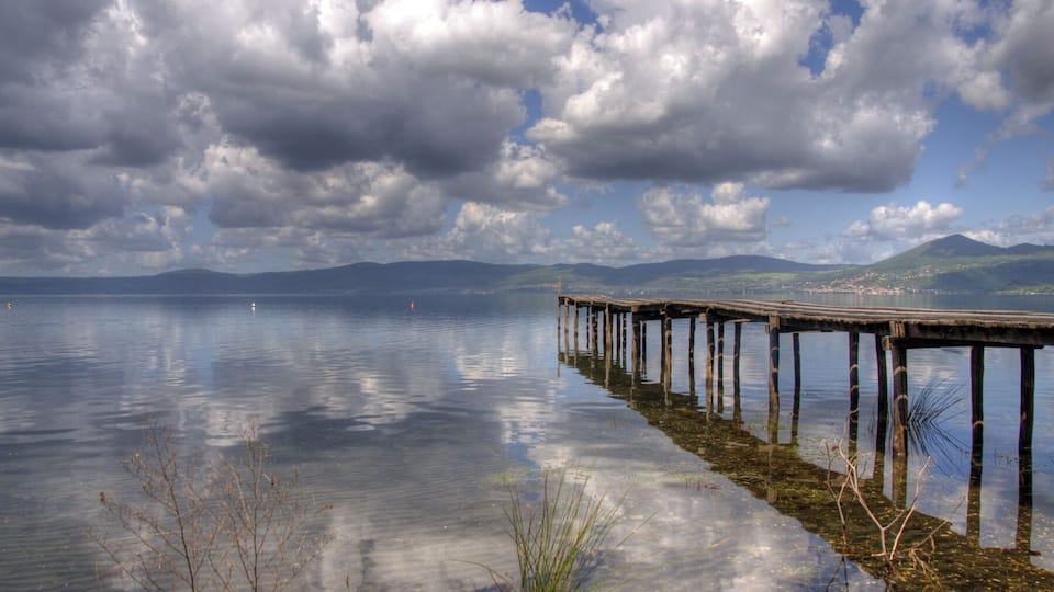A wooden pier extending into calm waters, surrounded by mountains and a partly cloudy sky.