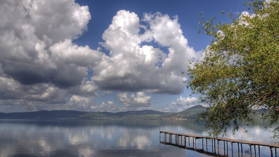 A calm lake with a wooden pier extending into the water, surrounded by lush greenery and a backdrop of mountains under a partly cloudy sky.