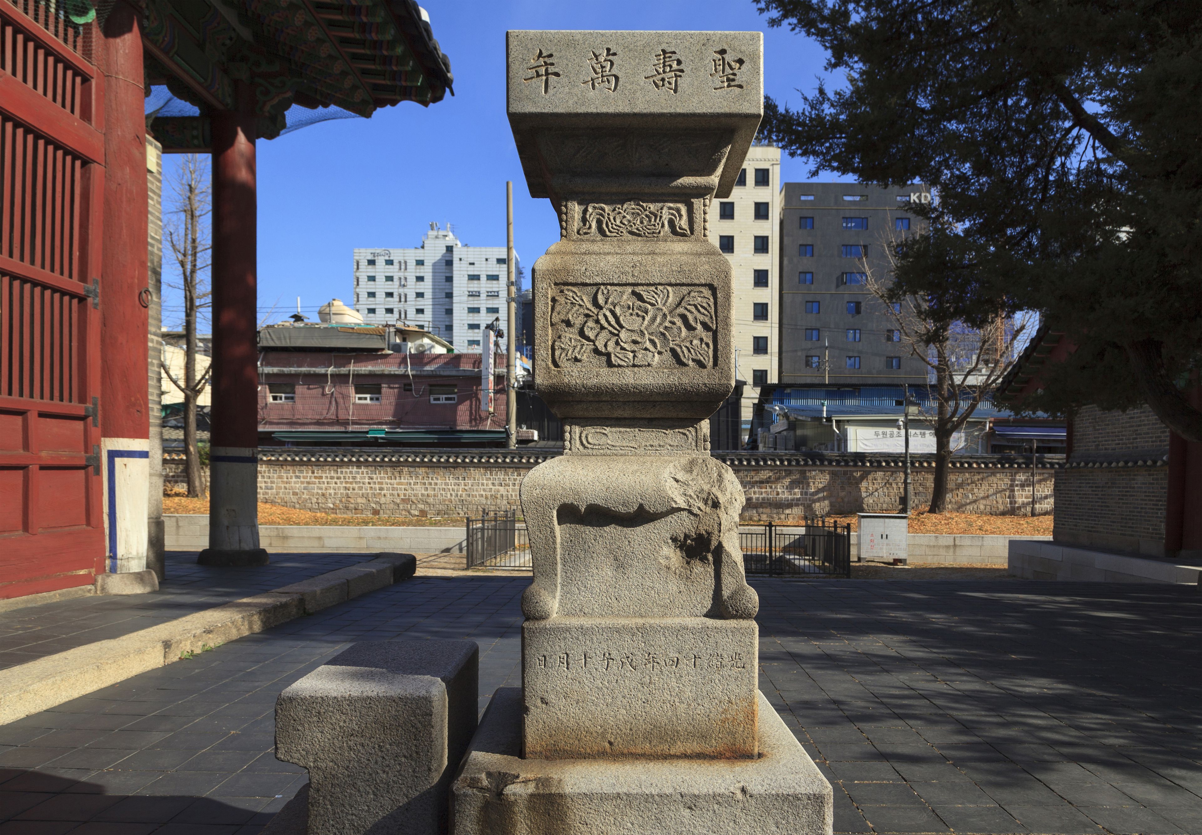 A stone monument with inscriptions, set against a backdrop of traditional architecture and modern buildings.
