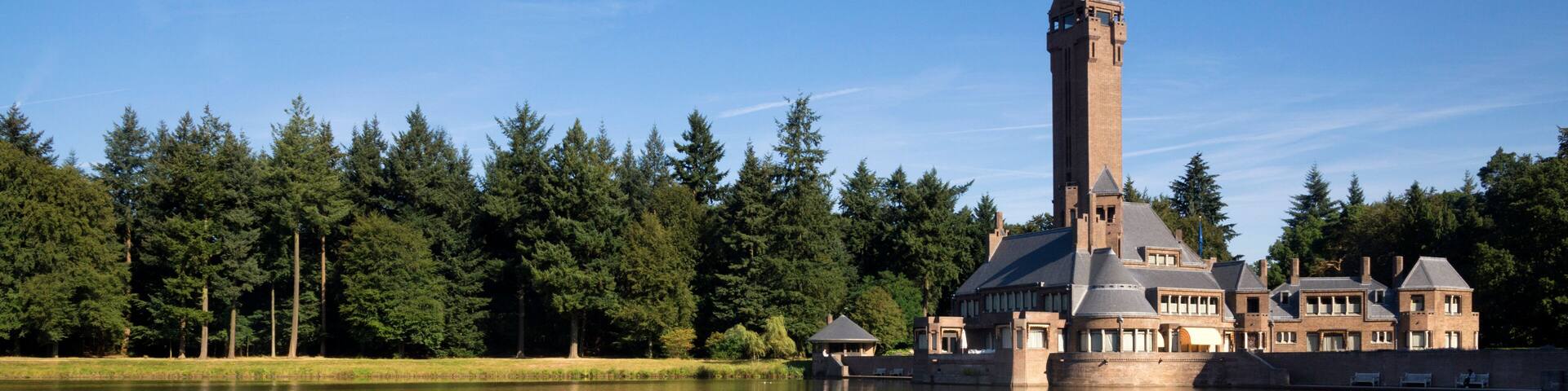 A large, historic building with a prominent tower, surrounded by a calm lake and dense forest.