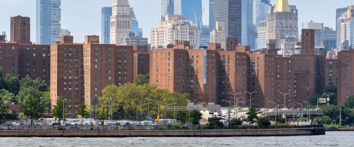 A city skyline with a mix of modern skyscrapers and historic buildings, a river in the foreground, and a clear sky.