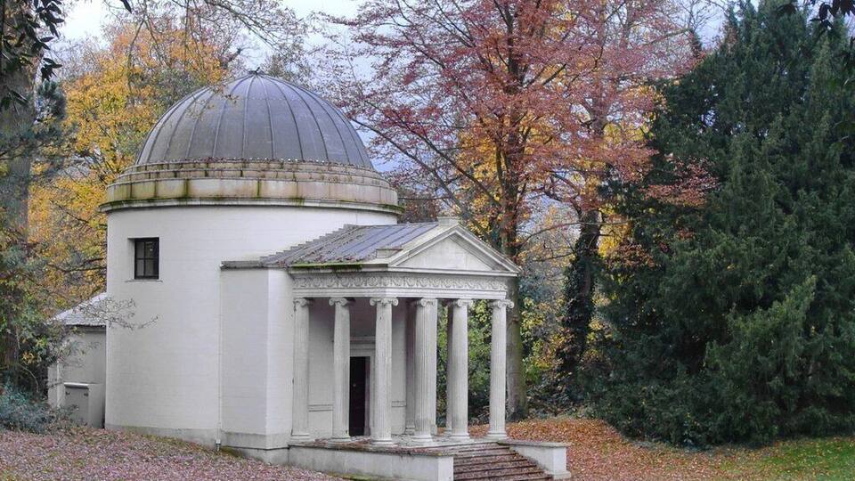 A small white building with a dome and columns, surrounded by trees with autumn foliage.
