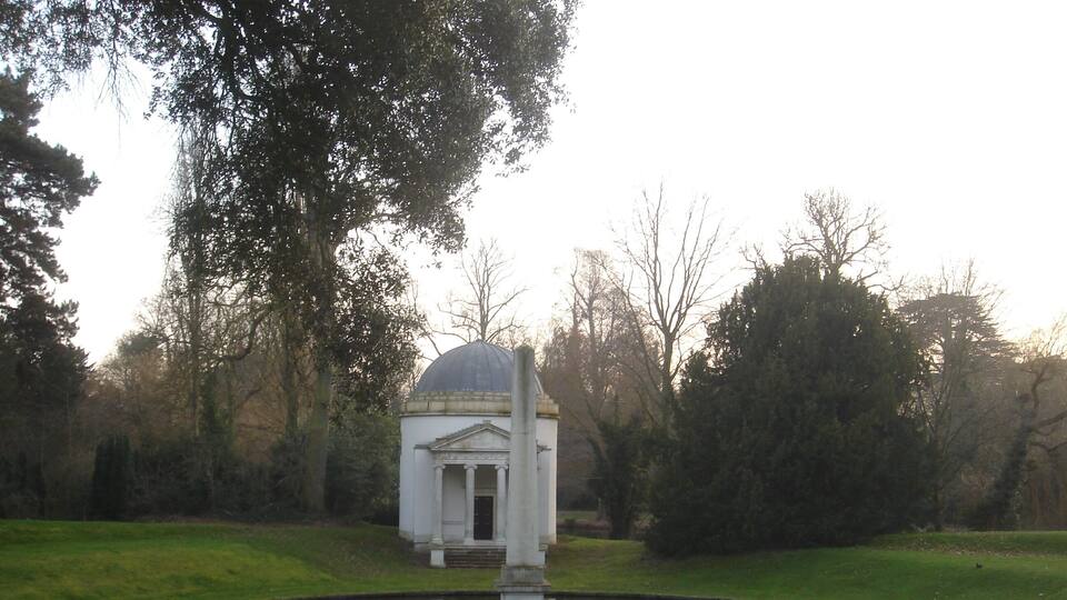 A small white building with a dome, surrounded by a pond and trees.