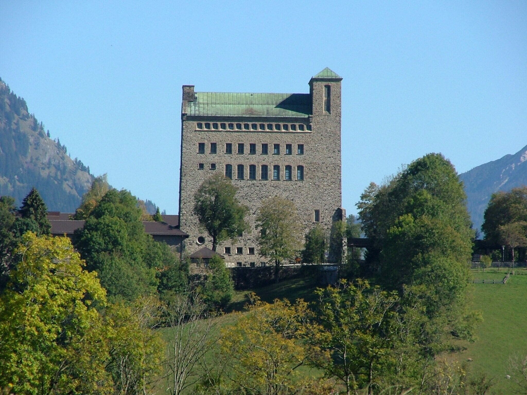 A stone tower with a green roof, surrounded by trees and mountains.