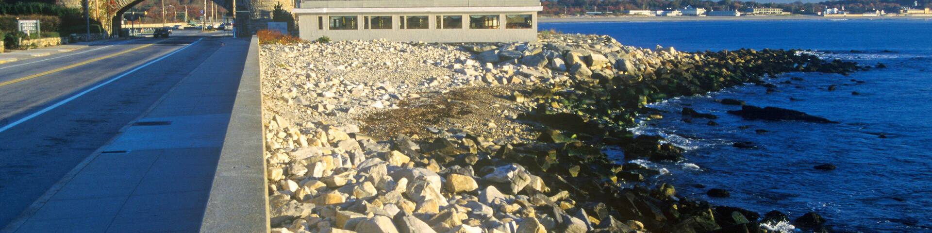 A coastal road with a stone bridge, a large building with a blue roof, and a rocky shoreline.