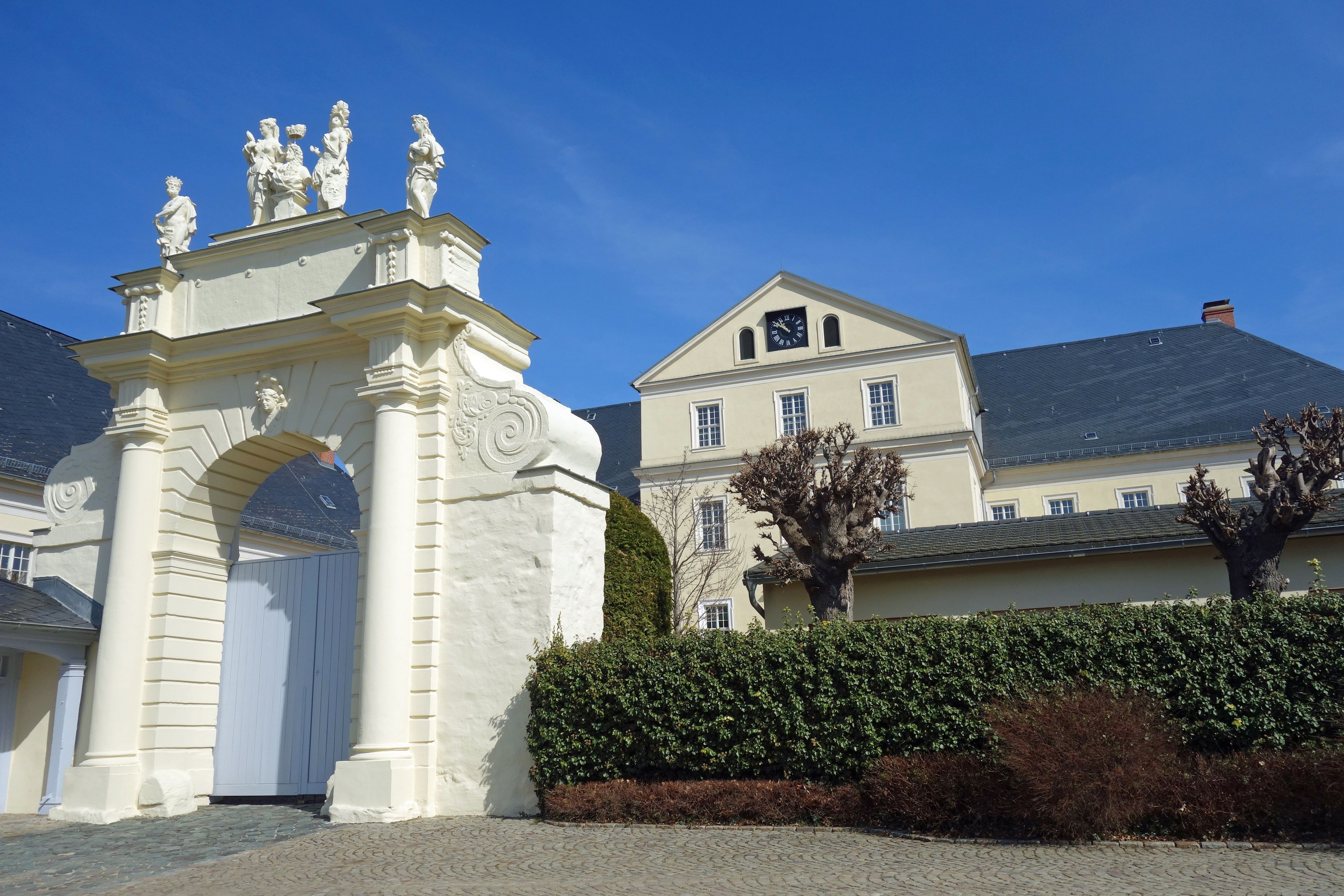 Ein klassisches weißes Tor mit Statuen, ein großes Gebäude mit einer Uhr und ein Baum im Vordergrund.