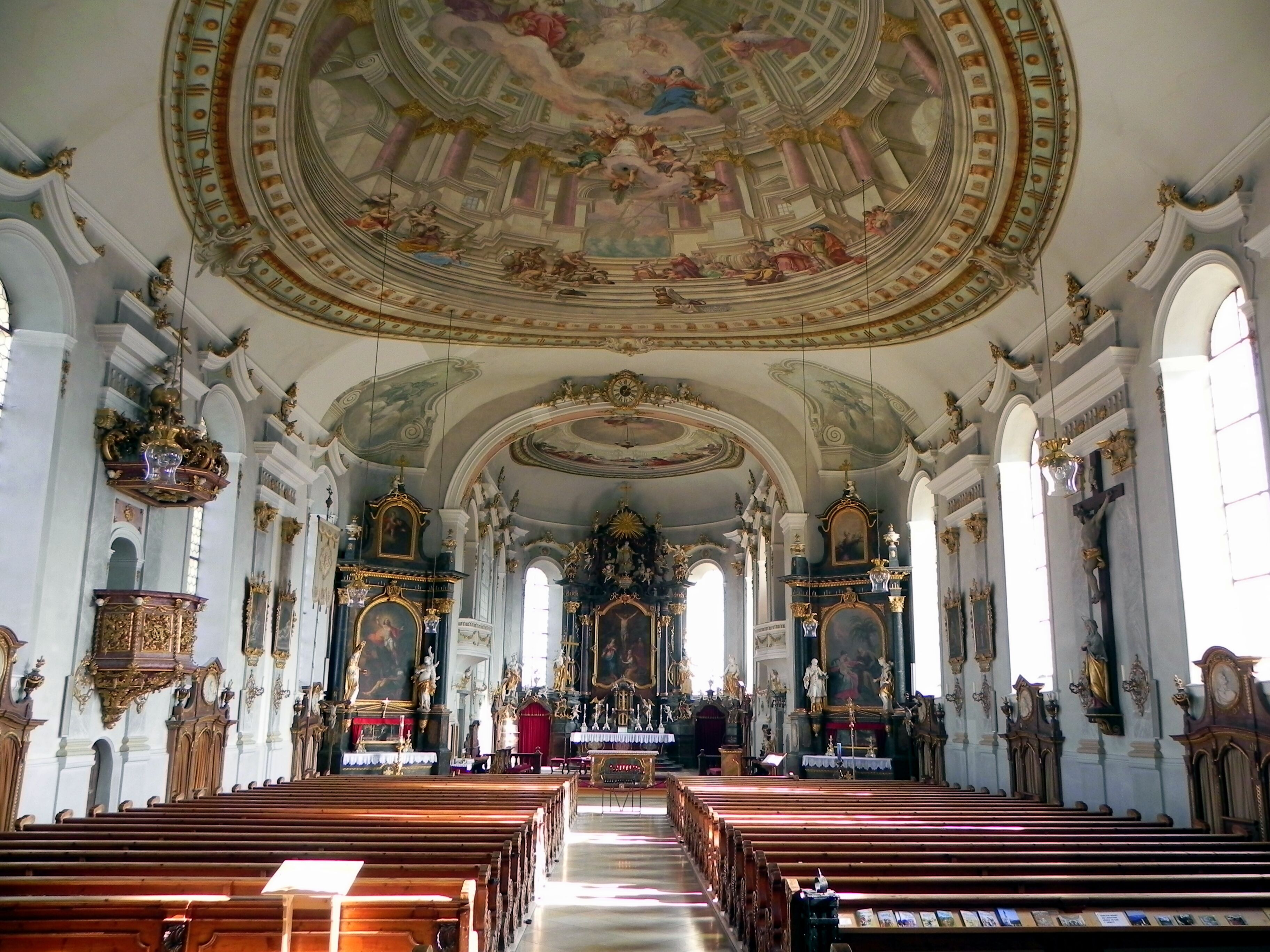 An ornate church interior with high ceilings, detailed frescoes, and rows of wooden pews.