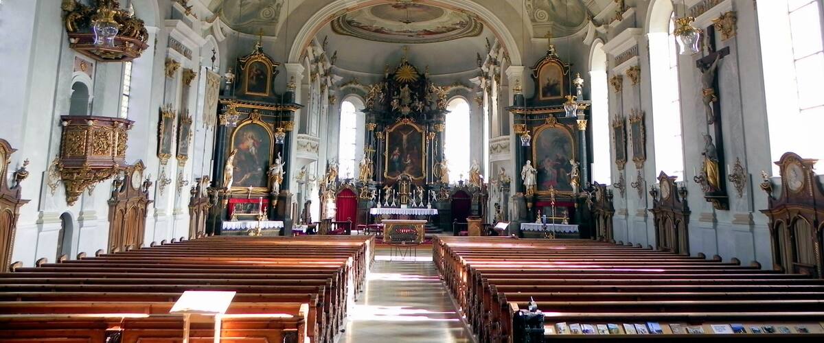 An ornate church interior with high ceilings, detailed frescoes, and rows of wooden pews.