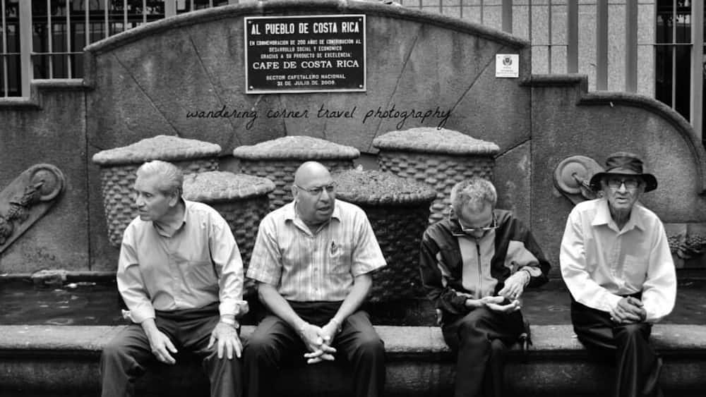 Quatre hommes sont assis sur un banc de pierre devant une fontaine, sur laquelle est apposée une plaque commémorative célébrant les 200 ans de contributions au développement social et économique du Costa Rica.