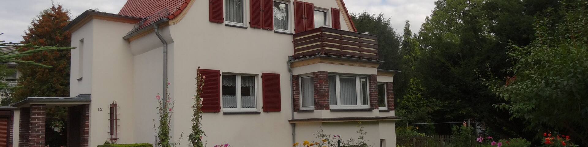 A two-story house with a red-tiled roof and red shutters, surrounded by a garden with various flowers and shrubs.