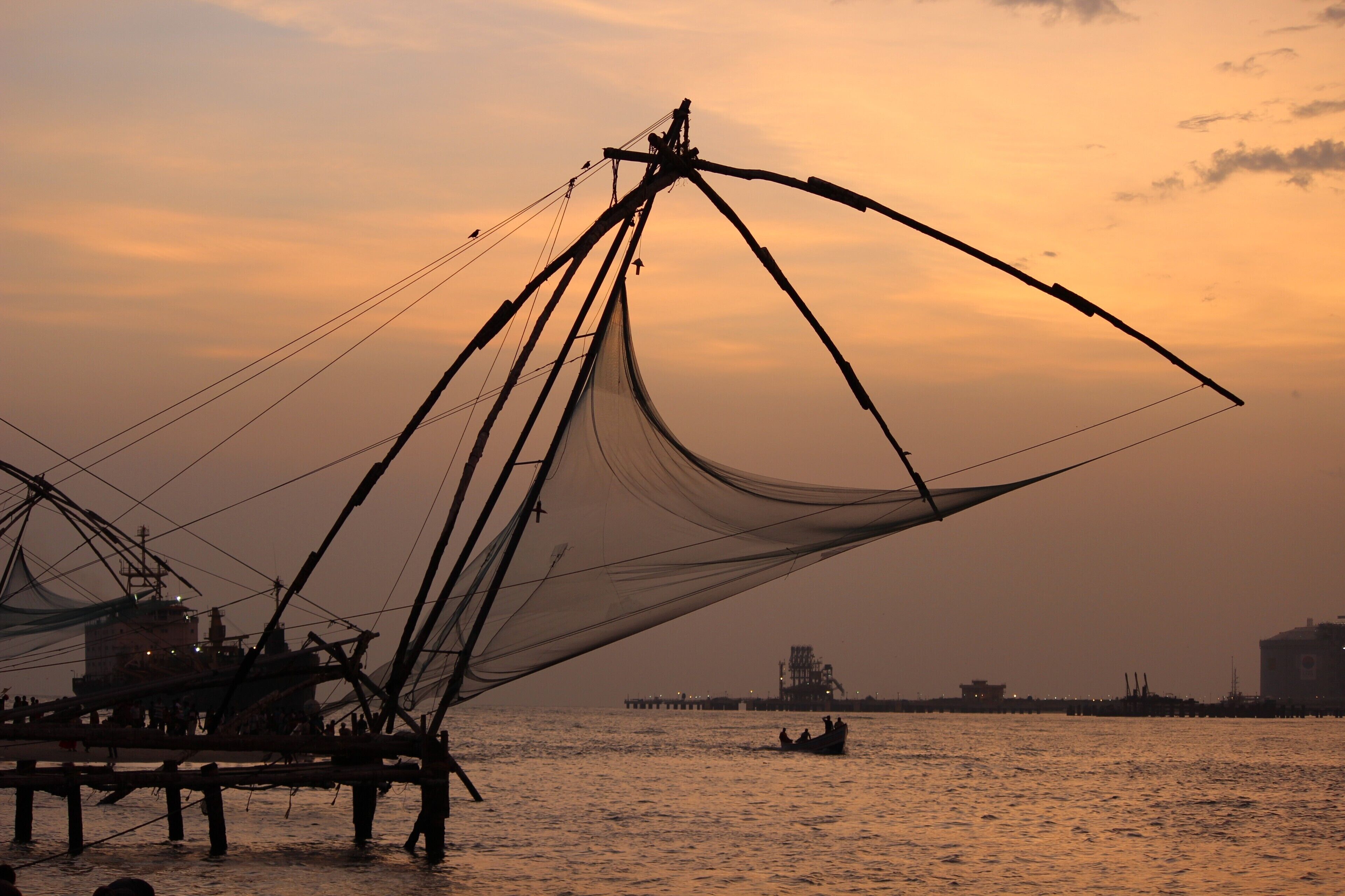 A fishing net structure at sunset with a boat in the water and a pier in the background.