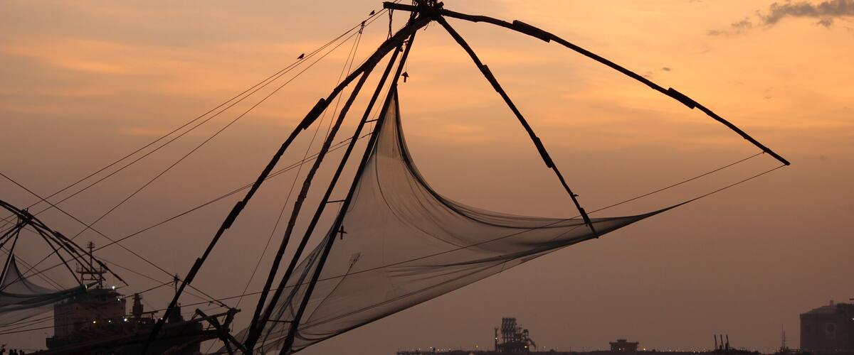 A fishing net structure at sunset with a boat in the water and a pier in the background.