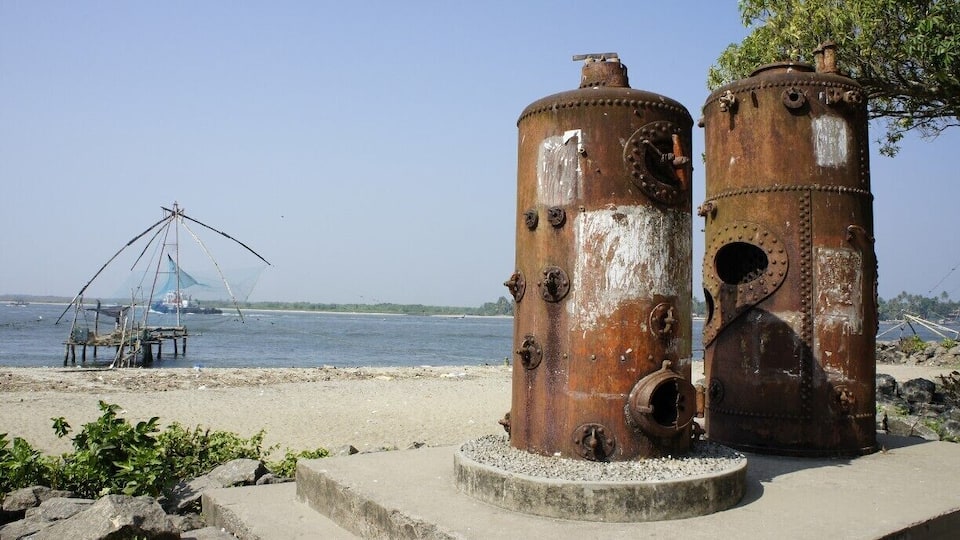 Two rusted cylindrical metal structures on a concrete platform by a body of water with a fishing net in the background.