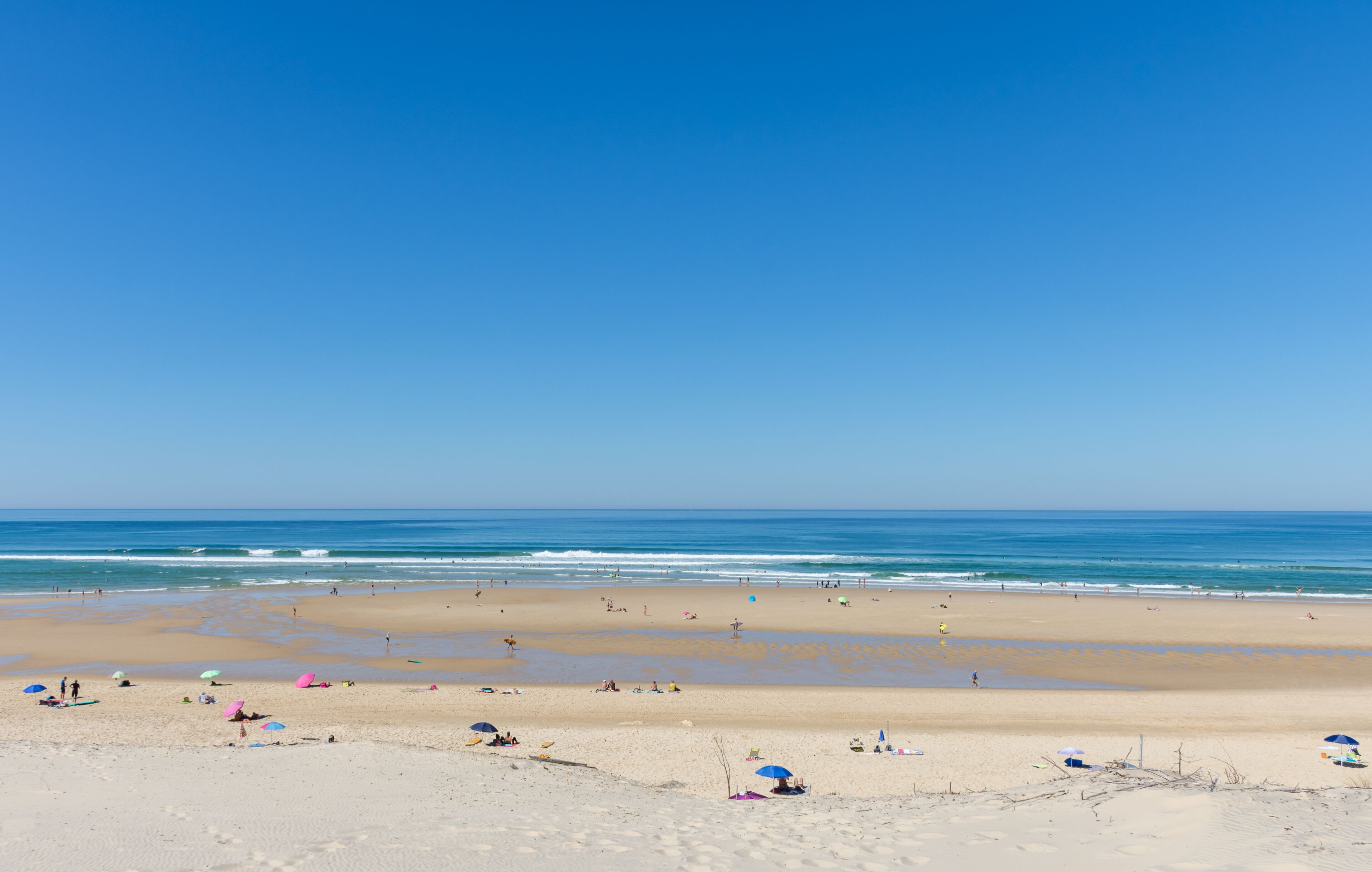 A wide sandy beach with gentle waves, clear blue sky, and a few umbrellas.