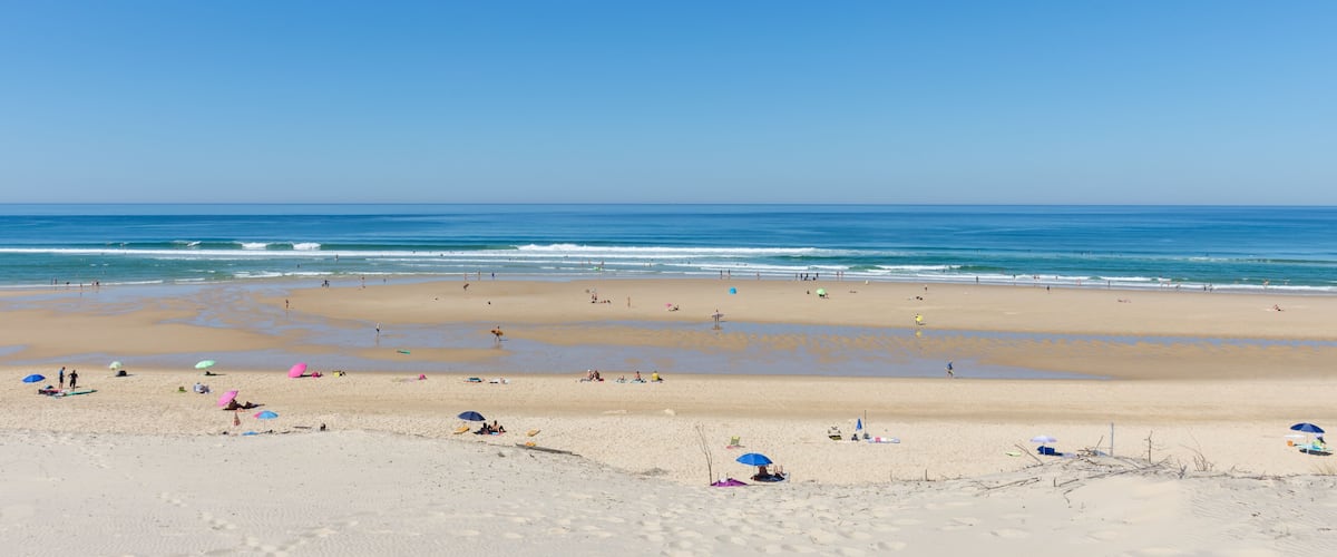 A wide sandy beach with gentle waves, clear blue sky, and a few umbrellas.