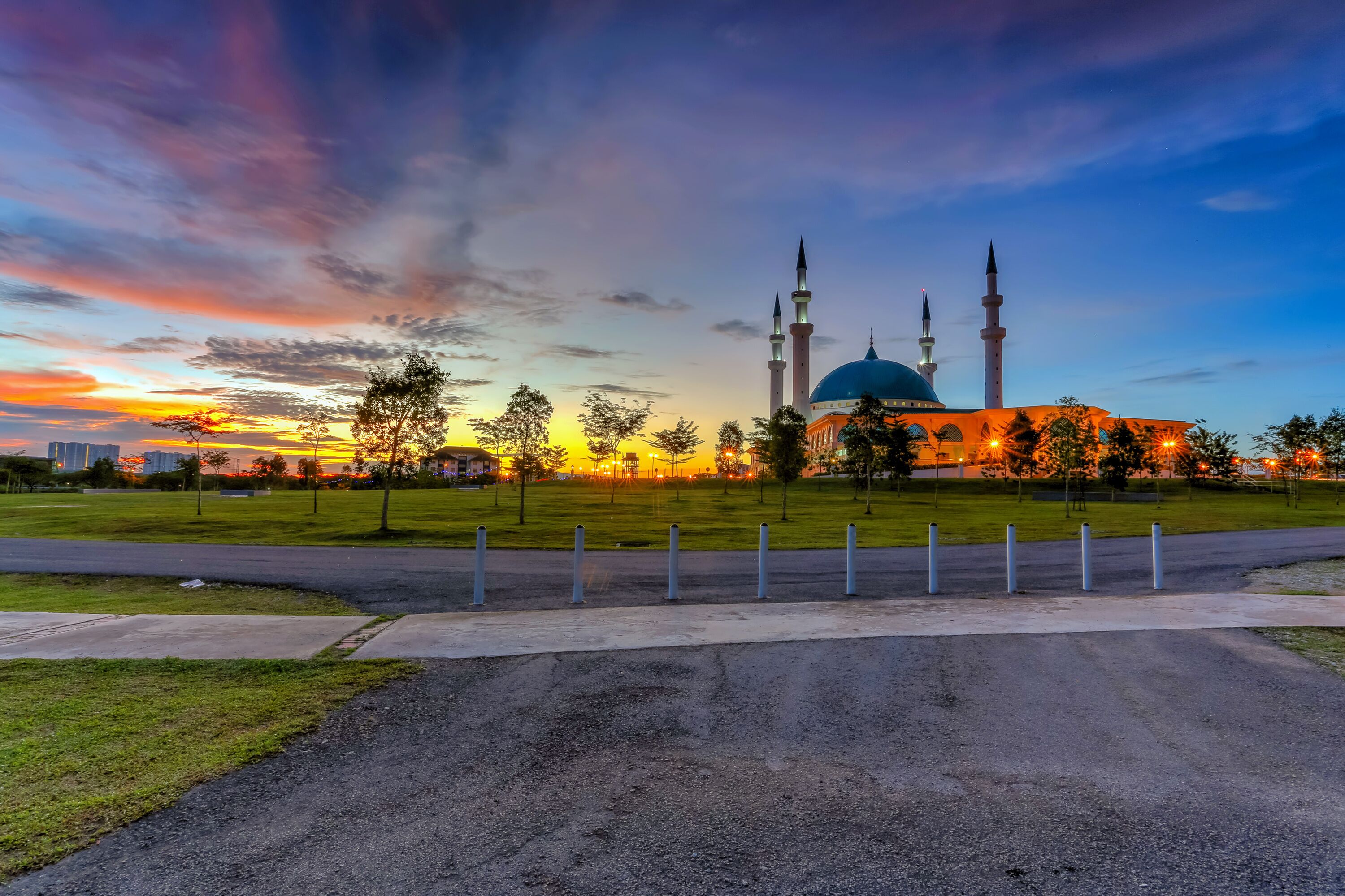 A mosque with a blue dome and three minarets, surrounded by a park with trees and a road.