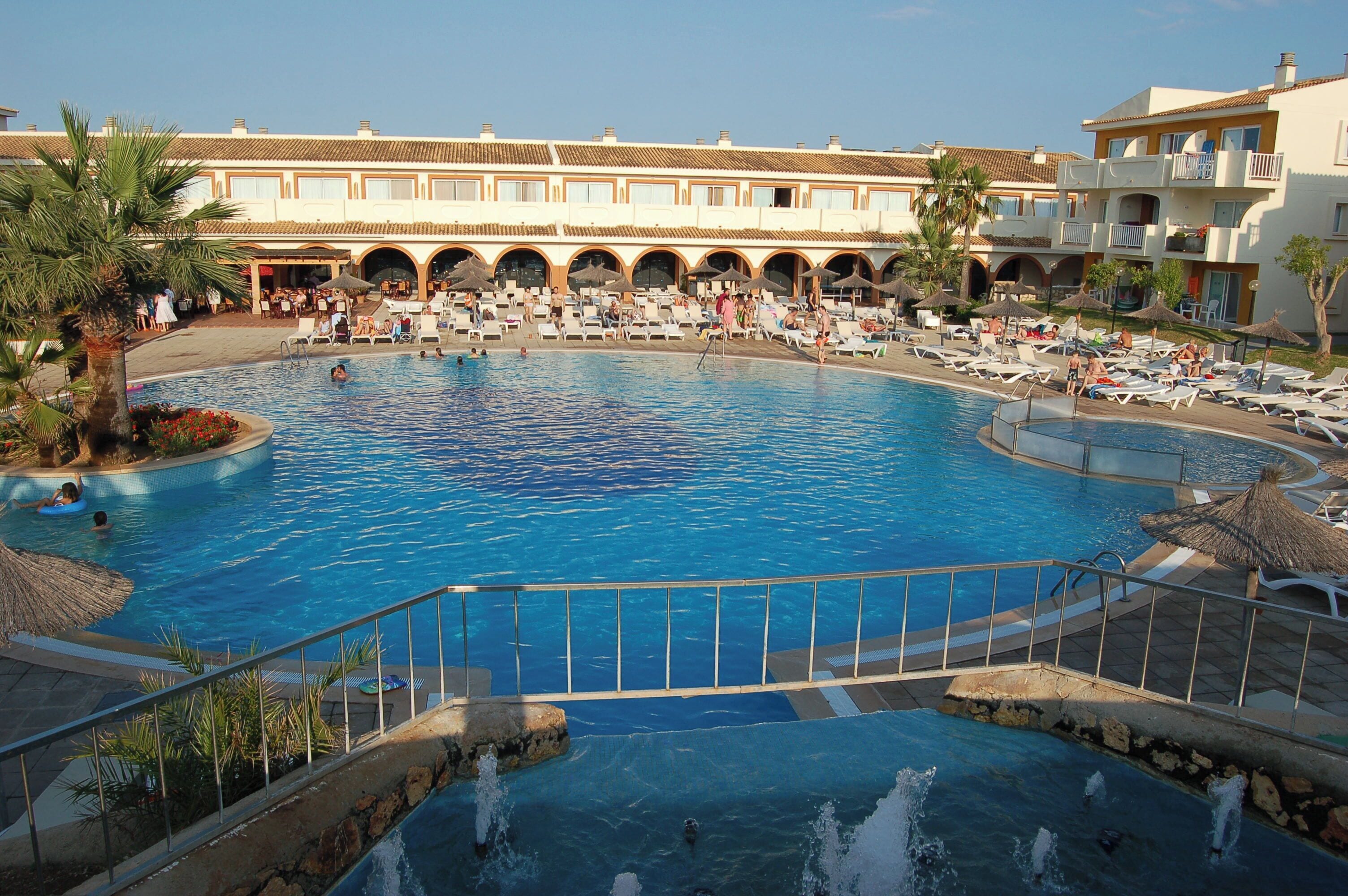 A large swimming pool with lounge chairs and umbrellas, surrounded by buildings with a Mediterranean architectural style.